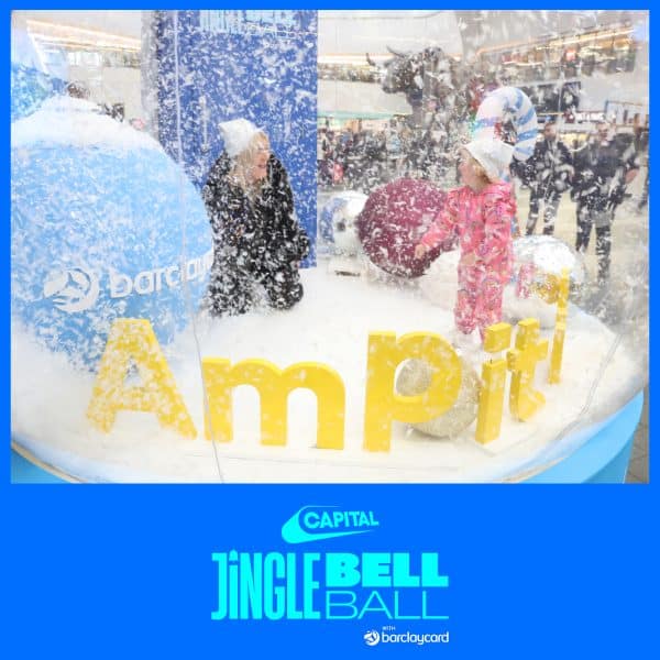 Two people are inside a snow globe-like display at the Capital Jingle Bell Ball event, surrounded by artificial snow and large colored balls.