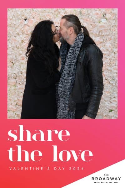 A couple kisses in front of a pink and white floral wall with text that reads "Share the Love - Valentine's Day 2024" and "The Broadway" at the bottom.