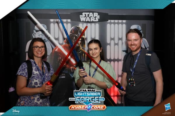 Three people holding lightsabers stand in front of a Star Wars backdrop with characters in the background.