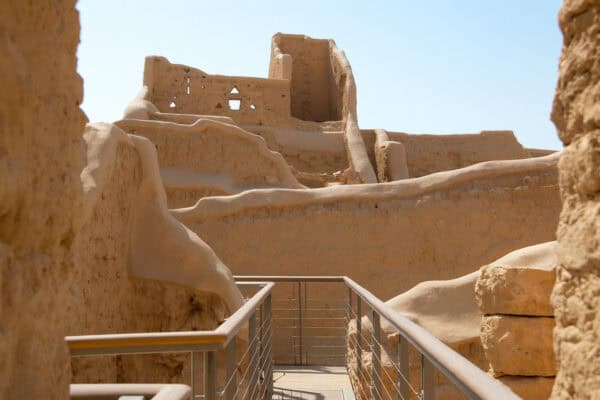 A metal walkway leads through ancient, sunlit mud-brick ruins with eroded walls and partially standing structures under a clear sky.