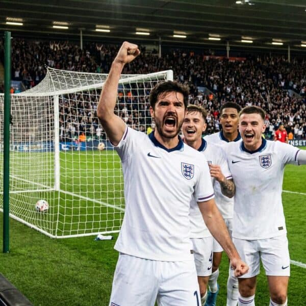 Four England football players celebrate near the goalpost, with one in the foreground raising his fist and shouting as a crowd cheers in the stadium behind them.