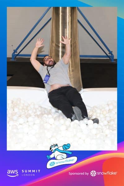 A man with glasses is sliding down into a ball pit filled with white balls. The event is sponsored by AWS Summit London and Snowflake, indicated by logos at the bottom of the image.